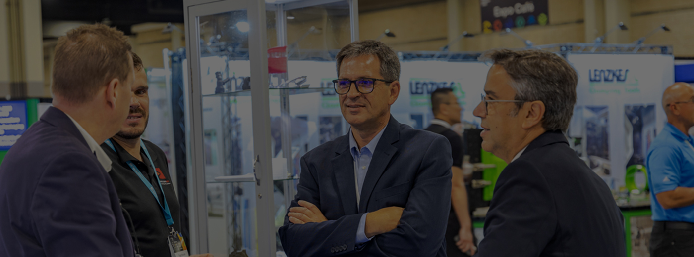 A diverse group of individuals engaged in conversation at a bustling trade show, surrounded by various displays and booths.