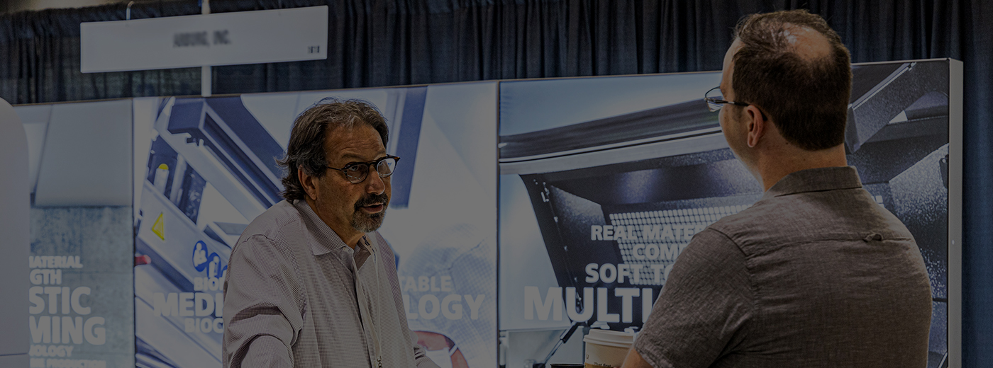 An exhibitor and an attendee engaged in a conversation at the PBS TX show, with a booth display visible in the background.