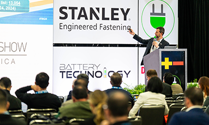 A man delivering a presentation at a conference, gesturing towards a large screen filled with visual content.