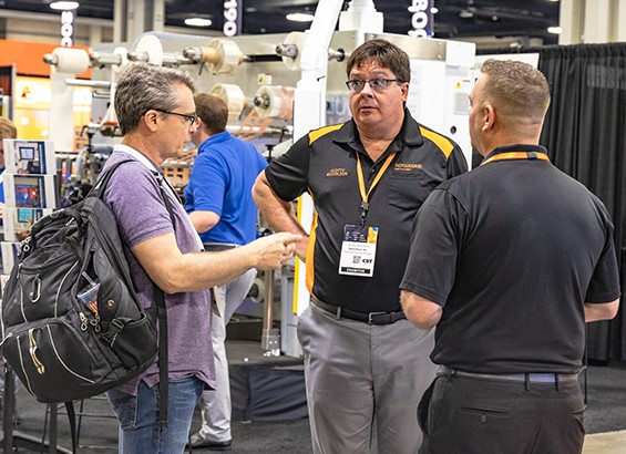 Three men engaged in conversation at a bustling trade show, surrounded by booths and attendees